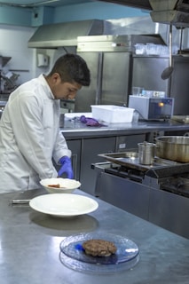 A chef in a white coat is preparing food in a commercial kitchen. He is wearing blue gloves and plating a dish on a stainless steel counter. The kitchen is equipped with industrial appliances including a stove and refrigerators.