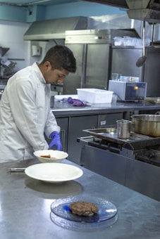 A chef in a white coat is preparing food in a commercial kitchen. He is wearing blue gloves and plating a dish on a stainless steel counter. The kitchen is equipped with industrial appliances including a stove and refrigerators.