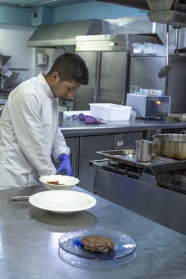 A chef in a white coat is preparing food in a commercial kitchen. He is wearing blue gloves and plating a dish on a stainless steel counter. The kitchen is equipped with industrial appliances including a stove and refrigerators.