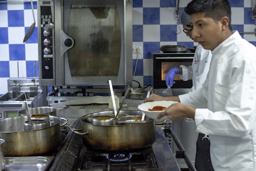 Two chefs are in a professional kitchen, engaged in cooking. One is wearing a white coat while holding a dish with red sauce, and another is in the background wearing a glove, appearing to work with the oven. The kitchen features large pots on the stove, metal appliances, and blue and white checkered tiles on the wall.