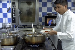 Two chefs are in a professional kitchen, engaged in cooking. One is wearing a white coat while holding a dish with red sauce, and another is in the background wearing a glove, appearing to work with the oven. The kitchen features large pots on the stove, metal appliances, and blue and white checkered tiles on the wall.