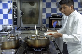 Two chefs are in a professional kitchen, engaged in cooking. One is wearing a white coat while holding a dish with red sauce, and another is in the background wearing a glove, appearing to work with the oven. The kitchen features large pots on the stove, metal appliances, and blue and white checkered tiles on the wall.
