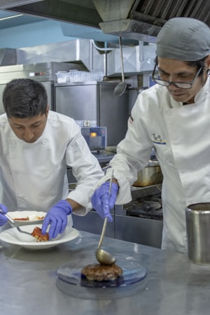 Two chefs are working in a professional kitchen, wearing white uniforms and blue gloves. One is plating food on a white dish, while the other carefully applies sauce to a cooked patty on a blue plate. The kitchen atmosphere includes industrial stainless steel equipment and a focused, busy scene.
