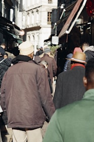 A bustling street market scene with people walking along narrow pathways between outdoor stalls. The individuals are wearing winter clothing, and the architecture in the background is European, with tall buildings and narrow windows.