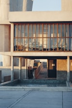 A modern architectural structure with large glass windows framed by concrete and wooden beams. The interior appears to be an office or meeting space with chairs and tables visible inside. The building is bathed in warm sunlight, casting long shadows across the exterior.