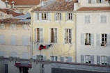 An old residential building with a yellow facade shows weathered and cracked paint. Laundry is hung on a line beneath a window where a person is slightly visible. The windows have shutters, and some have plants on the ledges. Roofs with reddish-brown tiles contrast with the lighter building colors.