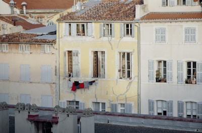 An old residential building with a yellow facade shows weathered and cracked paint. Laundry is hung on a line beneath a window where a person is slightly visible. The windows have shutters, and some have plants on the ledges. Roofs with reddish-brown tiles contrast with the lighter building colors.