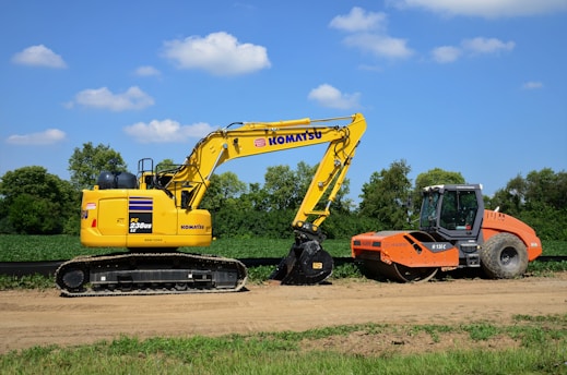 A yellow excavator and an orange road roller are positioned side by side on a dirt path with greenery and trees in the background under a clear blue sky.