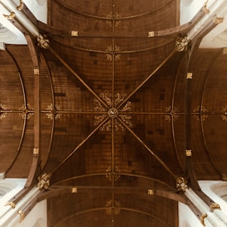 An intricate ceiling design featuring a central ornate wooden structure with gold embellishments. The ceiling is divided into sections by beams that form geometric shapes.
