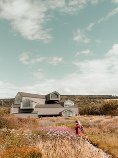 A futuristic building with angular, geometric design elements is surrounded by a colorful, blooming garden. The sky above is a clear blue with some scattered clouds. Several people are visible, walking through the garden.