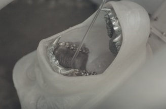 A close-up of a white sculpture resembling an open mouth with sharp, metallic teeth. The sculpture appears to be in a workshop or industrial setting, possibly undergoing a cleaning or crafting process.