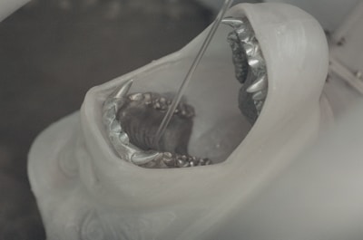 A close-up of a white sculpture resembling an open mouth with sharp, metallic teeth. The sculpture appears to be in a workshop or industrial setting, possibly undergoing a cleaning or crafting process.
