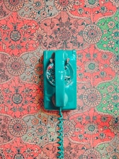 A vintage teal rotary dial telephone is placed on an intricately patterned, floral red and green wallpaper background. The vibrant, retro design contrasts with the bold color of the telephone, creating a striking visual.