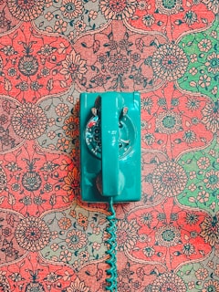 A vintage teal rotary dial telephone is placed on an intricately patterned, floral red and green wallpaper background. The vibrant, retro design contrasts with the bold color of the telephone, creating a striking visual.