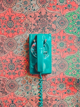 A vintage teal rotary dial telephone is placed on an intricately patterned, floral red and green wallpaper background. The vibrant, retro design contrasts with the bold color of the telephone, creating a striking visual.