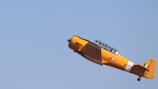 A bright yellow vintage airplane is flying at an upward angle in a clear blue sky. The aircraft features an Indian roundel and tail fin. Its design is reminiscent of mid-20th-century trainer planes, with a glazed cockpit canopy.