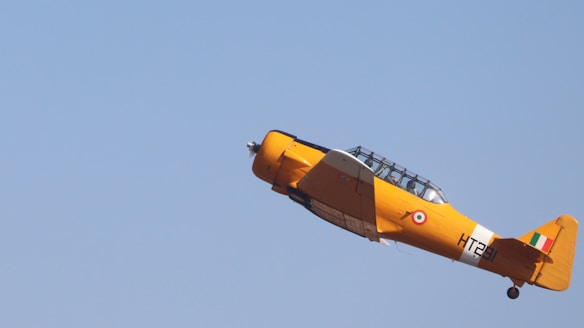 A bright yellow vintage airplane is flying at an upward angle in a clear blue sky. The aircraft features an Indian roundel and tail fin. Its design is reminiscent of mid-20th-century trainer planes, with a glazed cockpit canopy.