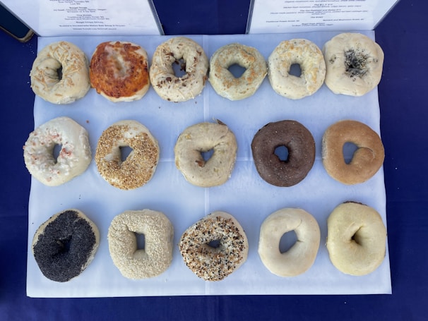 A display of various bagels arranged in rows on a white sheet. The bagels have different toppings and flavors, including poppy seeds, sesame seeds, and others. The background is a dark blue fabric, and menus or descriptions are partially visible at the top.