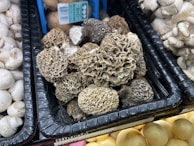 A display of various mushrooms at a market or store, with a central focus on a pile of morel mushrooms. The morels have a distinctive honeycomb-like texture and earthy brown color. Surrounding them are other types of mushrooms, such as white button and oyster mushrooms.