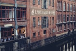 A brick building is reflected in the still waters of a canal. The structure has large red-framed windows and the sign 'E. Michaelis & Co' prominently displayed on the wall. Some windows are illuminated from within, suggesting interior activity. The building features classic architectural details, creating a historic and industrial feel.