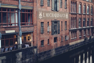 A brick building is reflected in the still waters of a canal. The structure has large red-framed windows and the sign 'E. Michaelis & Co' prominently displayed on the wall. Some windows are illuminated from within, suggesting interior activity. The building features classic architectural details, creating a historic and industrial feel.