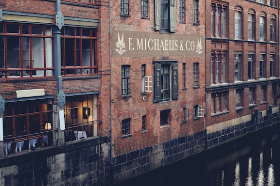 A brick building is reflected in the still waters of a canal. The structure has large red-framed windows and the sign 'E. Michaelis & Co' prominently displayed on the wall. Some windows are illuminated from within, suggesting interior activity. The building features classic architectural details, creating a historic and industrial feel.