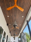 A modern outdoor ceiling features wooden panels with several ceiling fans and ornate hanging lanterns. The ceiling fans have wooden blades, and the lanterns exhibit intricate detailing. The space has a bright and airy atmosphere, with a glimpse of spiral stairs and green foliage outside the structure.