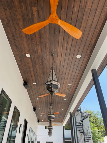 A modern outdoor ceiling features wooden panels with several ceiling fans and ornate hanging lanterns. The ceiling fans have wooden blades, and the lanterns exhibit intricate detailing. The space has a bright and airy atmosphere, with a glimpse of spiral stairs and green foliage outside the structure.