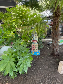A whimsical sculpture with a colorful, smiling face is situated among lush green foliage. The sculpture, painted with vibrant hues, features the words 'ART in Your Box' and is surrounded by various plants, including a palm and large-leafed greenery under a partially shaded area. A car is visible in the background, parked under a structure.