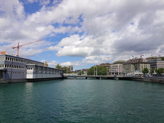 A river flows through a city with buildings lining both sides. A modern, gray building with a red crane is on the left, while more traditional architecture is visible on the right. A bridge spans across the river, with a tram moving over it. The sky is partially cloudy and the water is a calm green.