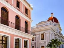 A street view showcasing colonial-style architecture with a pink building featuring arched windows and ornate balconies. Adjacent is a white building with intricate details and a prominent red dome. Bright sunlight enhances the scene, casting shadows against the pastel-colored facades.