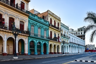 A row of colorful, colonial-style buildings line a quiet, empty street. The architecture features arched doorways and balconies with intricate railings. The colors of the facades range from cream and turquoise to pale yellow and white. A palm tree is visible on the right, adding an element of tropical scenery.