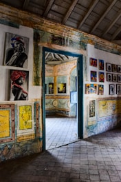 The image depicts an interior space, likely an art gallery or historic building, with colorful artwork adorning the walls. The room is decorated with vibrant paintings which include portraits and abstract designs. The architecture features patterned tiles on the floor and walls with a rustic door leading to another room, where more artwork is visible.