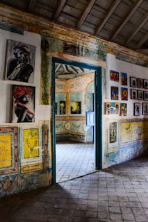The image depicts an interior space, likely an art gallery or historic building, with colorful artwork adorning the walls. The room is decorated with vibrant paintings which include portraits and abstract designs. The architecture features patterned tiles on the floor and walls with a rustic door leading to another room, where more artwork is visible.