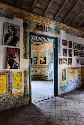 The image depicts an interior space, likely an art gallery or historic building, with colorful artwork adorning the walls. The room is decorated with vibrant paintings which include portraits and abstract designs. The architecture features patterned tiles on the floor and walls with a rustic door leading to another room, where more artwork is visible.