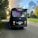 A black bus with a humorous mustache decoration on the front, parked on a road lined with trees. A colorful crown emblem is displayed above the windshield. Soft purple lighting is visible inside, against a backdrop of a partly cloudy sky with sunlight streaming through.