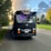 A black bus with a humorous mustache decoration on the front, parked on a road lined with trees. A colorful crown emblem is displayed above the windshield. Soft purple lighting is visible inside, against a backdrop of a partly cloudy sky with sunlight streaming through.