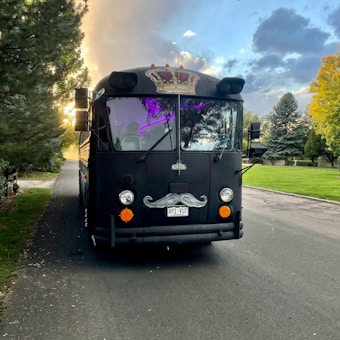 A black bus with a humorous mustache decoration on the front, parked on a road lined with trees. A colorful crown emblem is displayed above the windshield. Soft purple lighting is visible inside, against a backdrop of a partly cloudy sky with sunlight streaming through.