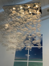 A modern art installation featuring numerous transparent glass orbs hanging from the ceiling creates an ethereal atmosphere. The orbs are spherical in shape and varied in size, with multiple small lights illuminating them from above. In the background, a large print split into four sections displays a serene seascape with a blue sky and scattered clouds.