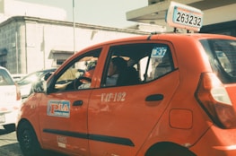 A red taxi with signage on the side and top is parked on a sunlit street. The taxi has a number plate with a series of numbers and letters displayed on top. In the background, there are other vehicles and a building with a stone facade.