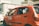 A red taxi with signage on the side and top is parked on a sunlit street. The taxi has a number plate with a series of numbers and letters displayed on top. In the background, there are other vehicles and a building with a stone facade.