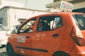 A red taxi with signage on the side and top is parked on a sunlit street. The taxi has a number plate with a series of numbers and letters displayed on top. In the background, there are other vehicles and a building with a stone facade.