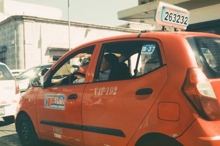 A red taxi with signage on the side and top is parked on a sunlit street. The taxi has a number plate with a series of numbers and letters displayed on top. In the background, there are other vehicles and a building with a stone facade.