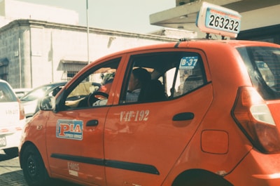 A red taxi with signage on the side and top is parked on a sunlit street. The taxi has a number plate with a series of numbers and letters displayed on top. In the background, there are other vehicles and a building with a stone facade.