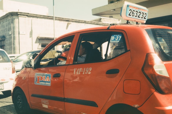 A red taxi with signage on the side and top is parked on a sunlit street. The taxi has a number plate with a series of numbers and letters displayed on top. In the background, there are other vehicles and a building with a stone facade.