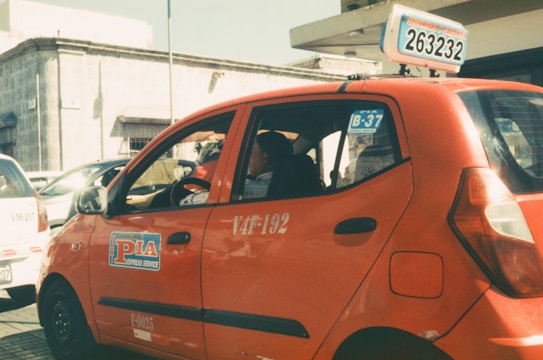A red taxi with signage on the side and top is parked on a sunlit street. The taxi has a number plate with a series of numbers and letters displayed on top. In the background, there are other vehicles and a building with a stone facade.