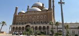 A grand historical mosque with multiple domes and minarets, showcasing Islamic architecture. The structure is built with brown stone and features arched windows and doorways. In the foreground, there are palm trees and a well-maintained garden. A group of people is visible, possibly tourists, walking towards the entrance.
