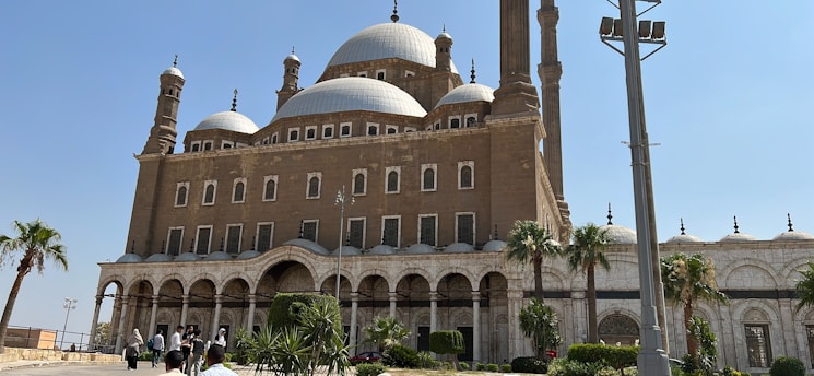 A grand historical mosque with multiple domes and minarets, showcasing Islamic architecture. The structure is built with brown stone and features arched windows and doorways. In the foreground, there are palm trees and a well-maintained garden. A group of people is visible, possibly tourists, walking towards the entrance.