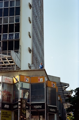 A modern building with a mostly glass facade and beige concrete elements is prominently featured. Multiple commercial signs including 'Hâthi' and 'Atelier das Unhas' are visible on the lower floors. High-rise architecture with a grid of reflective windows extends upward. A person in a blue outfit stands on an upper ledge, looking out.