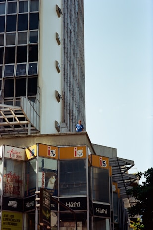 A modern building with a mostly glass facade and beige concrete elements is prominently featured. Multiple commercial signs including 'Hâthi' and 'Atelier das Unhas' are visible on the lower floors. High-rise architecture with a grid of reflective windows extends upward. A person in a blue outfit stands on an upper ledge, looking out.